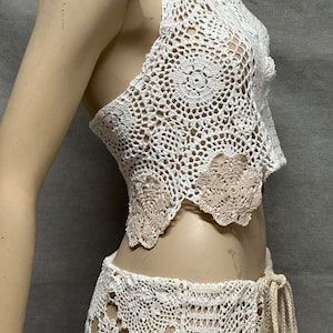 Puede incluir: Un conjunto de top y falda de crochet blanco. El top tiene un dobladillo festoneado y la falda tiene una cintura con cordón.