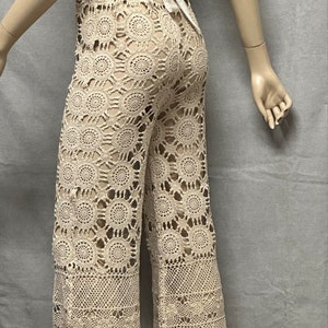 May include: Beige crochet trousers with a wide leg and a scalloped hem. The trousers are made of a delicate, open-weave fabric.