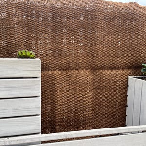 Op de afbeelding: Twee witte houten plantenbakken met planten die erin groeien. De plantenbakken staan voor een bruine geweven wandbekleding.