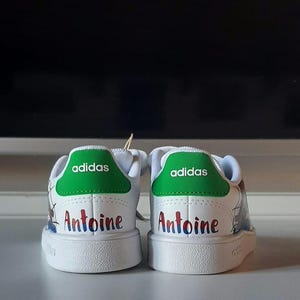 May include: Pair of white Adidas sneakers with green heel tabs. The word "adidas" is printed in white on the green tab. The name "Antoine" is written in colorful letters on the back of each shoe. The shoes are on a white surface.