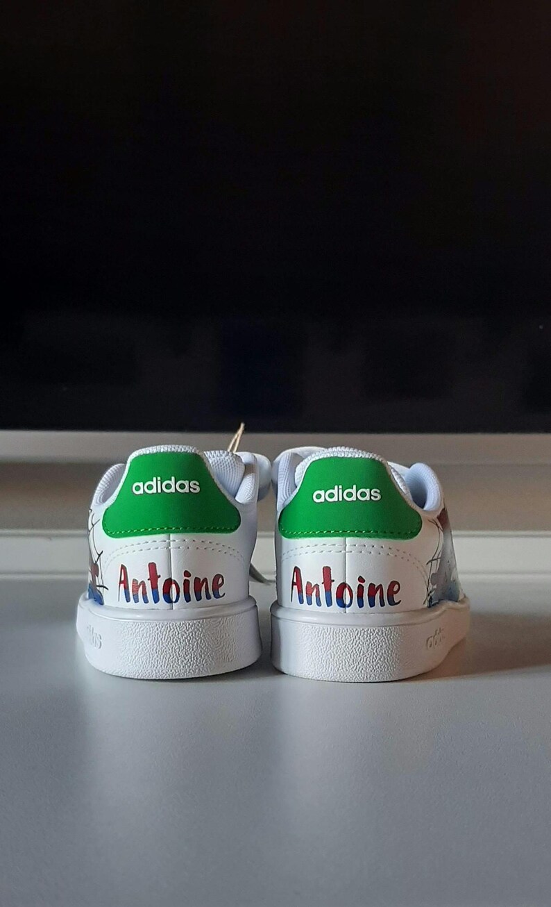 May include: Pair of white Adidas sneakers with green heel tabs. The word "adidas" is printed in white on the green tab. The name "Antoine" is written in colorful letters on the back of each shoe. The shoes are on a white surface.