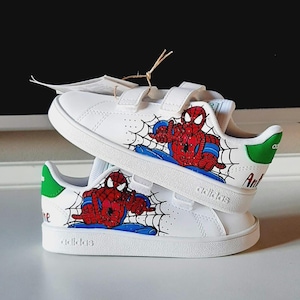 May include: White sneakers with hook-and-loop straps, featuring a Spider-Man design. The shoes have a red and blue Spider-Man graphic with a spiderweb design. The heel has a green accent with the word "Ant" printed on the side.
