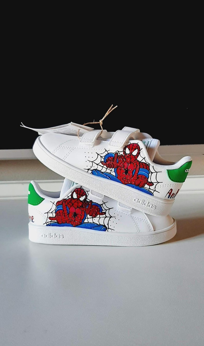 May include: White sneakers with hook-and-loop straps, featuring a Spider-Man design. The shoes have a red and blue Spider-Man graphic with a spiderweb design. The heel has a green accent with the word "Ant" printed on the side.
