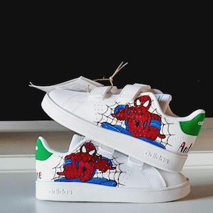 May include: White sneakers featuring a Spiderman design. The shoes have a red and blue Spiderman graphic on the side, with a black web design. The heel has a green accent. The shoes have velcro straps.