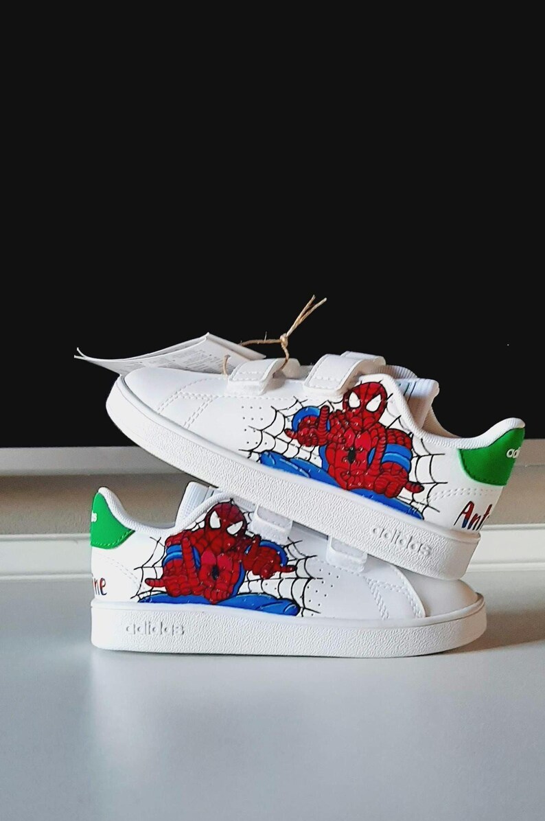 May include: White sneakers featuring a Spiderman design. The shoes have a red and blue Spiderman graphic on the side, with a black web design. The heel has a green accent. The shoes have velcro straps.