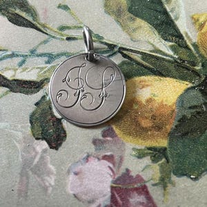 May include: A silver charm with a circular design featuring an engraved monogram of the letters "FP" in a decorative script.