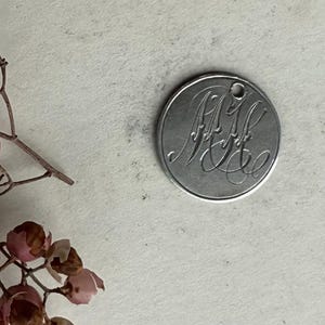 May include: A silver-colored, round pendant with an engraved monogram. The pendant has a small hole at the top for hanging. The monogram is in an elegant, cursive font. The background is a neutral, off-white color.