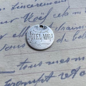 Puede incluir: Una ficha de metal de color plateado con la palabra "ADEL AIDA" inscrita en ella. La ficha es circular y tiene un borde ligeramente elevado.