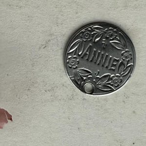 以下が含まれることがあります： 中央に「ANNIE」という名前が刻まれた、銀色の丸いペンダント。ペンダントは葉と花で構成された花の模様で囲まれています。ペンダントの下部には小さな穴があります。