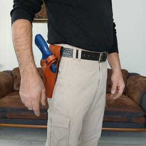 Könnte beinhalten: Ein braunes Lederholster mit einer blauen Schusswaffe ist an einem schwarzen Gürtel befestigt. Der Gürtel wird mit beigefarbenen Cargohosen getragen. Die Person trägt ein schwarzes Langarmshirt.
