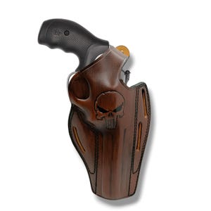 Pode incluir: Uma coldre de couro castanho com um emblema de caveira, segurando um revólver preto. O coldre tem um design detalhado com costuras visíveis e uma superfície texturizada. A empunhadura do revólver é preta, contrastando com a cor castanha do coldre.