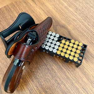 May include: A brown leather holster containing a handgun with a wooden grip, positioned next to a black plastic ammunition tray holding a mix of silver-colored and brass-colored cartridges arranged in a grid pattern on a wooden surface.