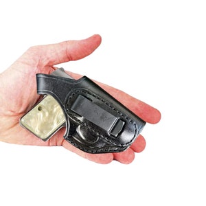 Puede incluir: Funda de cuero negro con un clip metálico, que sostiene una pequeña pistola con una empuñadura de color crema. La pistola es parcialmente visible dentro de la funda. La funda está diseñada para transporte oculto.