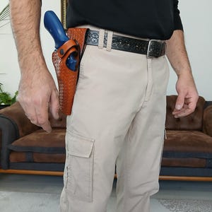 May include: A brown leather holster with a basketweave pattern holds a blue revolver. The holster is attached to a black belt with a silver buckle. The person is wearing khaki cargo pants and a black long-sleeve shirt.
