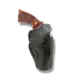 May include: A revolver with a polished brown wooden grip featuring a gold-colored circular emblem, partially inserted into a black leather holster. The holster displays a detailed basket-weave embossed pattern and visible black stitching along the edges.