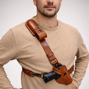 May include: A brown leather shoulder holster with a matching magazine pouch. The holster is designed to carry a handgun and is secured with a strap and buckle system. The leather has a rich, natural colour.