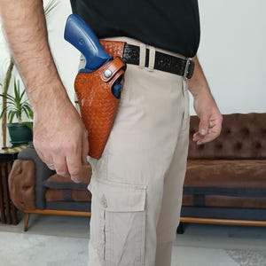 Peut inclure: Un étui en cuir beige avec un motif tressé et une fermeture à pression en métal argenté, contenant une arme d'entraînement bleue. L'étui est fixé à une ceinture en cuir noir à motifs portée sur un pantalon cargo beige et une chemise noire.