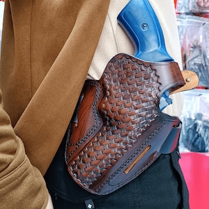 May include: A brown leather holster with a woven design, holding a blue handgun. The holster is worn on a black belt, with a brown jacket in the background. The holster is made of brown leather.