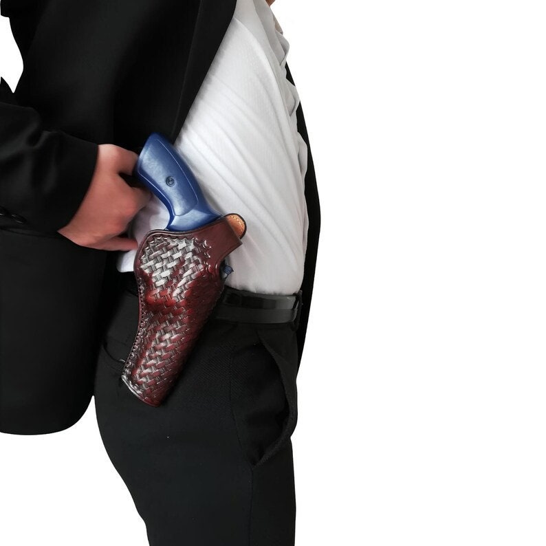 May include: A burgundy leather holster with a woven pattern, holding a blue handgun. The holster is worn on the hip, partially concealed under a black blazer and white shirt. The image is set against a white background.