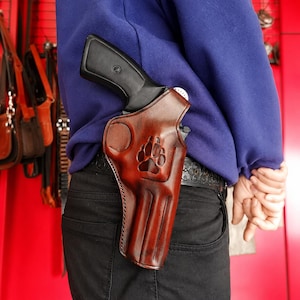 May include: A brown leather holster featuring a cutout paw print design, worn on a black belt. The holster contains a black handgun with a textured grip. The item is positioned against a blue sweatshirt and black trousers in a workshop setting.