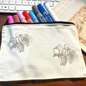 May include: A cream-colored canvas pencil case with a black zipper, filled with colorful markers. The case features a gray floral design. A keyboard and a beige pouch with a cat illustration are in the background.