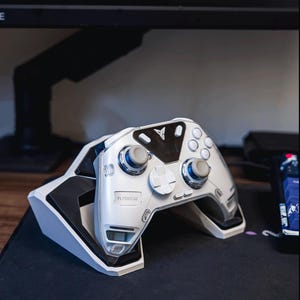 Könnte beinhalten: Ein weiß-silberner Gaming-Controller mit blauen Akzenten, der auf einer weiß-schwarzen Ladestation liegt. Der Controller hat ein schlankes Design mit mehreren Tasten und Joysticks. Der Markenname "FLYDIGI" ist auf dem Controller sichtbar.