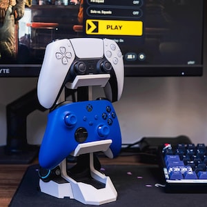 Könnte beinhalten: Ein weiß-blauer Gaming-Controller-Ständer, der einen weißen PlayStation 5-Controller und einen blauen Xbox-Controller hält. Der Ständer steht auf einem Schreibtisch mit einer Tastatur und einem Monitor, der eine Videospieloberfläche anzeigt. Der Monitor zeigt eine "PLAY"-Taste.