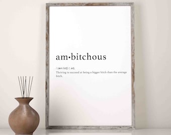 Op de afbeelding: Een ingelijste print met het woord "am·bitchous" in zwarte tekst. De definitie hieronder luidt: "Groeien om succesvoller te zijn dan de gemiddelde bitch." Een bruine vaas met zwarte stokken staat links.