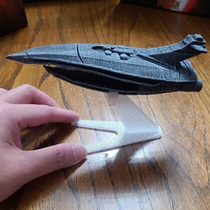 Puede incluir: Un modelo de nave espacial gris impreso en 3D en un soporte de plástico transparente. La nave espacial tiene un diseño elegante y futurista con una nariz puntiaguda y un cuerpo largo y delgado.