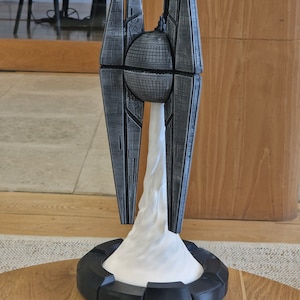 Könnte beinhalten: Eine futuristische, abstrakte Skulptur mit dunkelgrauer, strukturierter Oberfläche. Die Skulptur weist ein kugelförmiges Element und vertikale, architektonische Strukturen auf. Eine weiße, wolkenartige Form erhebt sich von einem schwarzen, kreisförmigen Sockel. Die Skulptur steht auf einer Holzoberfläche.
