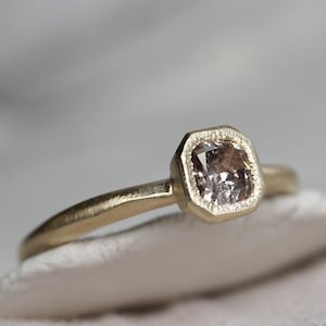 May include: A gold ring with a square-shaped diamond set in a bezel setting. The ring is on a white fabric background.