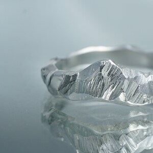 May include: A silver ring with a textured, mountain-like design. The ring is reflected in a shiny surface.