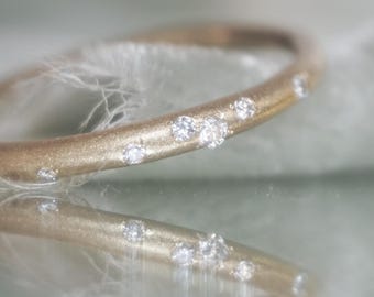 May include: A gold ring featuring small, sparkling gemstones set along the band. The ring is shown on a reflective surface, with its reflection below. The ring's design is simple and elegant, suitable for everyday wear.