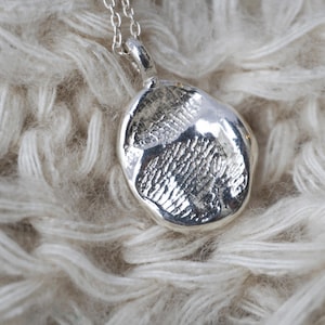 Op de afbeelding: Een zilveren ketting met een ronde, getextureerde hanger met een gedetailleerde afdruk van een poot.