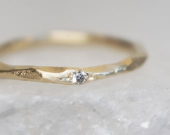 May include: A close-up of a gold ring with a small, clear gemstone. The ring has a textured, organic design. The gemstone is set in a prong setting. The ring is on a white surface.