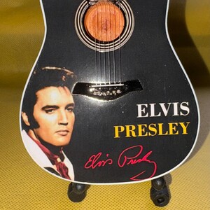 May include: Miniature black acoustic guitar with a portrait of Elvis Presley and his signature. The guitar has a wooden soundhole and strings. The text "ELVIS PRESLEY" is displayed in yellow and white.
