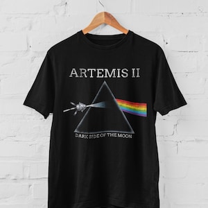 May include: Black t-shirt featuring the text "ARTEMIS II" above a graphic of a prism refracting light into a rainbow. Below the prism is the text "DARK SIDE OF THE MOON". The shirt is hanging on a wooden hanger.