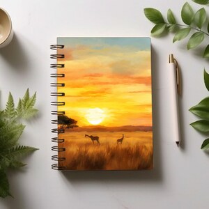 May include: A spiral bound notebook with a sunset scene on the cover. The scene features a golden sky with clouds, a setting sun, and two giraffes in a field of tall grass.