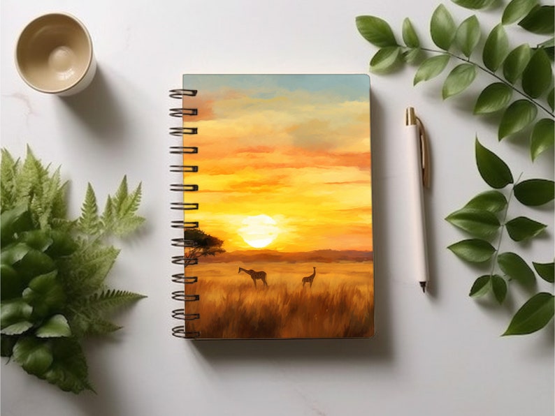 May include: A spiral bound notebook with a sunset scene on the cover. The scene features a golden sky with clouds, a setting sun, and two giraffes in a field of tall grass.