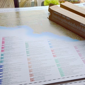 May include: A map of Los Angeles with a list of murders investigated by Lieutenant Columbo. The map is printed on a white background with blue, green, and red markers. The map is rolled up and sitting on a table next to a brown cardboard tube with text in French and English.