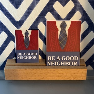 May include: Two rectangular cards with the text "BE A GOOD NEIGHBOR." are displayed on a wooden holder. The cards feature a graphic of a red sweater, a white collared shirt, and a plaid tie. The background has a blue and white geometric pattern.