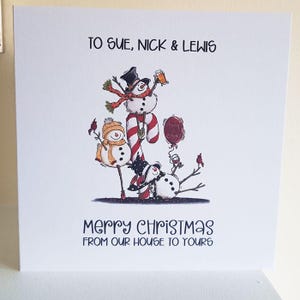 May include: A white Christmas card with a whimsical illustration of snowmen. The top text reads "TO SUE, NICK & LEWIS." The bottom text says "MERRY CHRISTMAS FROM OUR HOUSE TO YOURS." The snowmen are wearing hats and scarves.