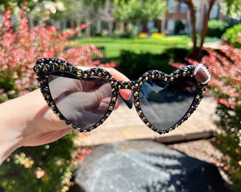 May include: Black heart-shaped sunglasses with gold accents and rhinestones. The sunglasses have the word "SAINTS" spelled out in black beads on the left lens and a small football charm on the right lens.