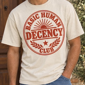 Może przedstawiać: Kremowy t-shirt z krótkim rękawem z okrągłą grafiką w kolorze rdzawego pomarańczu na klatce piersiowej. Wzór zawiera ikonę wschodzącego słońca nad napisem "BASIC HUMAN DECENCY CLUB" z gałązkami laurowymi i małą gwiazdką na dole.