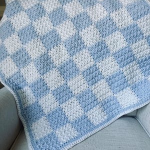 May include: A handmade, crocheted blanket in a checkerboard pattern of light blue and white. The blanket is draped over a light gray armchair. The edges are finished with a white border.