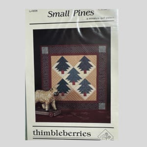 May include: A miniature quilt pattern titled "Small Pines" by thimbleberries. The pattern features a central design of five stylized evergreen trees in shades of blue and green, set against a tan and red background. A decorative sheep figurine is in the foreground.