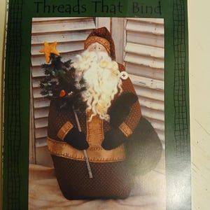 Op de afbeelding: Een stoffen kerstmanpop met een lange witte baard, die een kleine kerstboom met een ster vasthoudt. De pop draagt een rode hoed en jas met bruine accenten. De tekst "Threads That Bind" staat bovenaan.