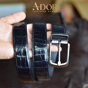 Puede incluir: Un cinturón negro con textura de piel de cocodrilo brillante y una hebilla de metal oscuro mate. El cinturón presenta costuras visibles a lo largo de los bordes y lleva el texto ADOR LEATHER STORE en la parte superior central.