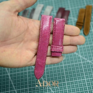 May include: A vibrant fuchsia pink watch strap featuring a textured lizard-skin pattern. The two-piece strap consists of a long section and a shorter buckle section with a sliding loop. The brand name ADOR LEATHER STORE is printed in gold at the bottom.
