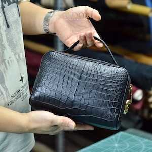May include: A black rectangular handbag with a crocodile skin texture. The bag has a small handle and a gold-coloured zip. The bag is held by a person.